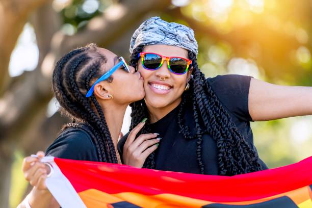 afro-latinx giovane lesbica che bacia sua moglie con in mano uno striscione arcobaleno con la parola proud scritta su di esso - couple black american culture kissing foto e immagini stock