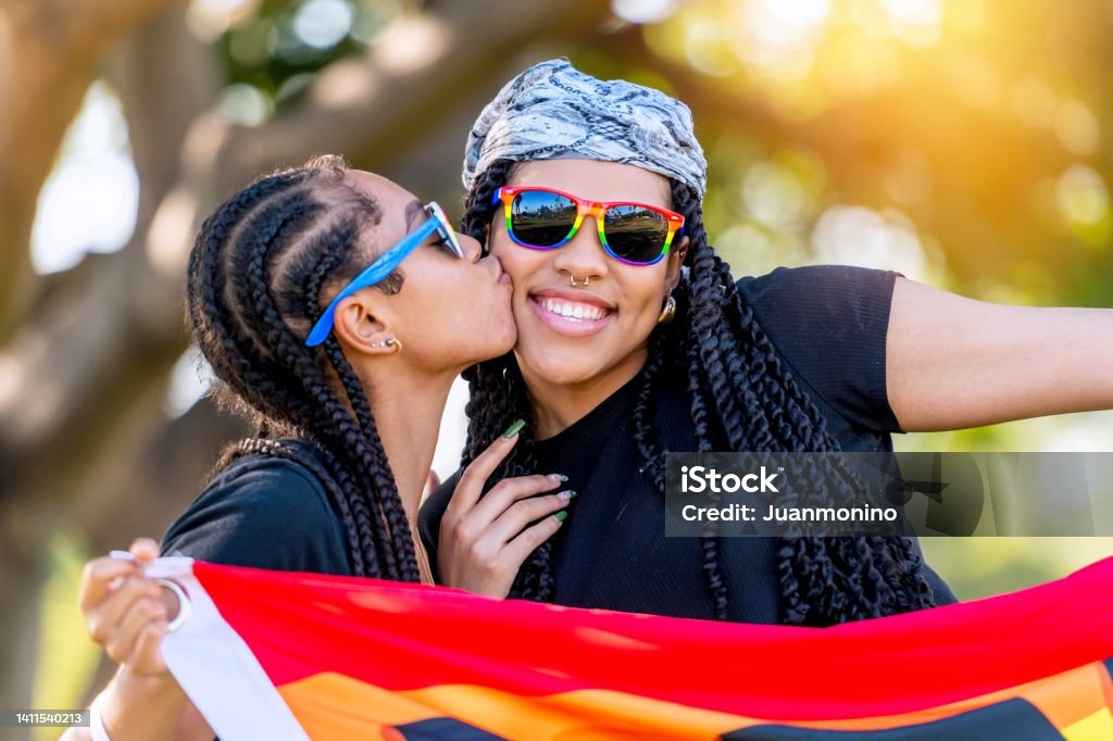 Afro-latinx young lesbian kissing her wife holding a rainbow banner with the word Proud written on it Kissing Stock Photo