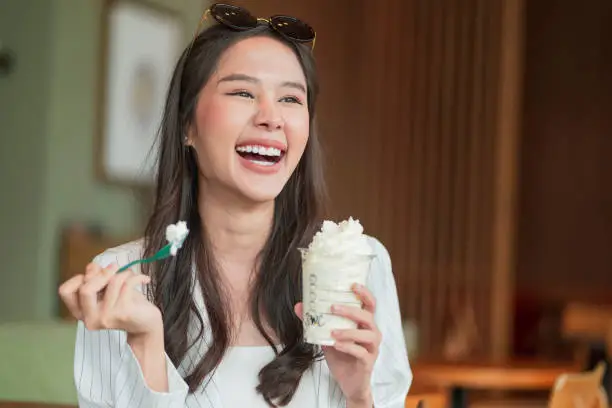 Photo of happiness joyful asian young adult female woman teasing yummy smiling hand hold whipped cream in plastic cup,enjoy eating asia woman toothy smile while take a break with glass of dessert in cafe