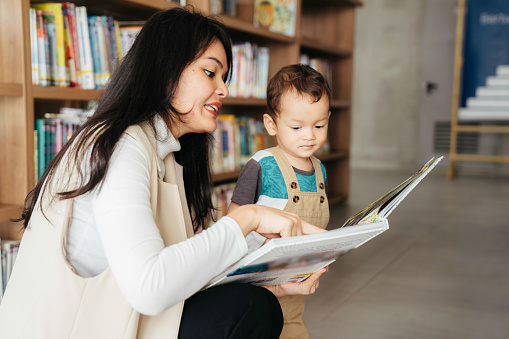 Young mother reading books to lovely little son in library