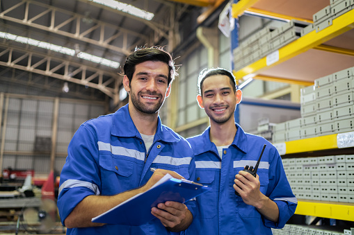 Portrait of two male industrial worker working in manufacturing plant. Young handsome male industry factory engineer wear helmet and processes order and product at manufactory warehouse with happiness