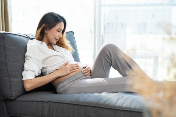 joven mujer asiática embarazada sosteniendo barriga con ambas manos sentada en el sofá de casa. la niña embarazada mira el estómago. tacto de la sensación del bebé dentro del vientre. pensando en que el recién nacido nazca en el futuro - animal uterus audio fotografías e imágenes de stock