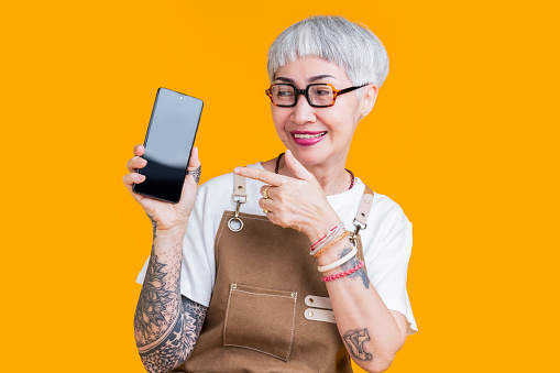 greeting old senior asian female business owner in apron hand gesture show smartphone blank screen with joyful presentation,asia woman tattoo wear glasses barista hand pointing  show cellphone online