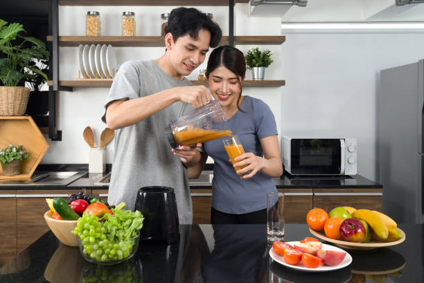 young man in casual clothe pour mixed fruit and veggie smoothie from blending machine into his girlfriend glass. asian lover spend  morning time together in the kitchen. - blender apple banana color image imagens e fotografias de stock