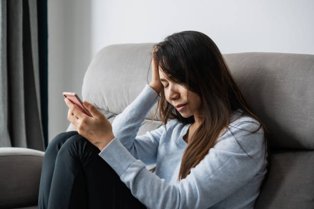 istock Stressed young Asian woman suffering on depression while holding mobile phone on hand sitting on couch in living room at home. Sad, unhappy, disappointed concept. Close up, copy space 1411548385