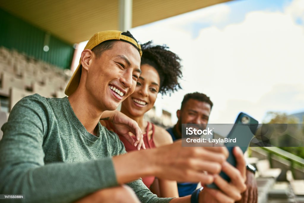 People outdoors smiling at phone, dressed casually. Young, diverse group looking and laughing at website on mobile excitedly. Friends sitting with each other on stadium chairs Three people outdoors smiling at phone. Two men and one woman are on stadium chairs. The group has gotten approval to take part in a marathon, while resting after training they found out. Friendship Stock Photo