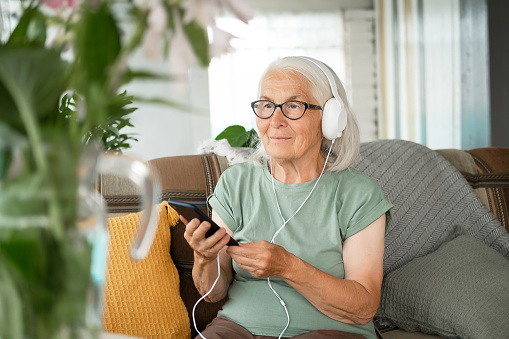 Caucasian elderly gray haired woman with headphones is sitting at home on couch with smartphone and listening to music, audiobook, lecture. Concept of audio healing and sound therapy, learning