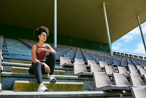 A happy female athlete resting on the stadium stair after a good workout. An active and fit woman taking a break outdoors on the training ground. An athletic lady happy about her fitness routine