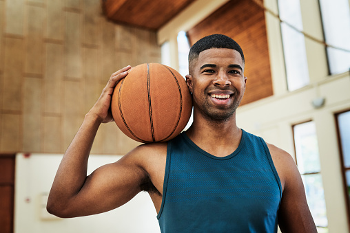A Happy African American basketball player holding a ball in a sports club. Portrait of a fit and active athlete enjoying his sports training at the college courts. Athletic man smiling at practice