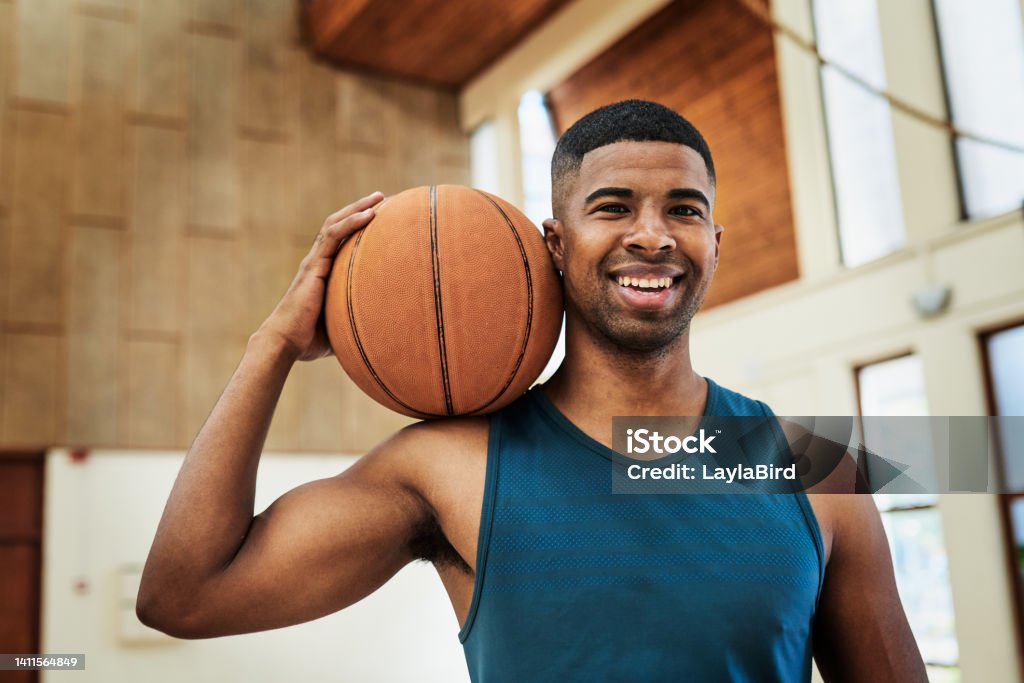 A Happy African American basketball player holding a ball in a sports club. Portrait of a fit and active athlete enjoying his sports training at the college courts. Athletic man smiling at practice Basketball - Ball Stock Photo