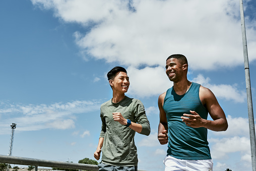 Active men jogging outdoors on blue cloudy sky with copy space. Two athletic guys or young sports friends running together, doing their routine cardio workout and fitness exercise in the city