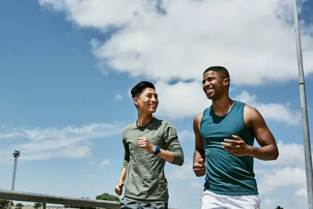 Photo of Active men jogging outdoors on blue cloudy sky with copy space. Two athletic guys or young sports friends running together, doing their routine cardio workout and fitness exercise in the city