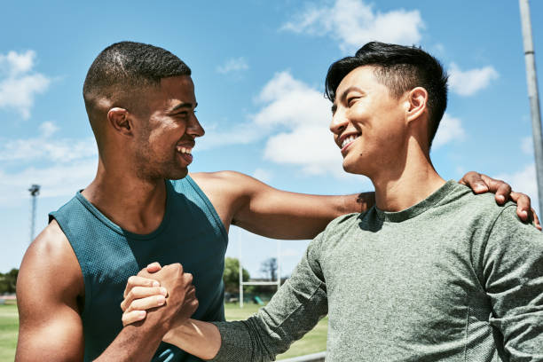 Motivated athletes giving a handshake and hugging in a friendly greeting on a sport field outside on a sunny day. Two young male friends celebrating victory and embracing in joy after winning a race stock photo