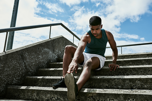 African athlete stretching during his workout on stairs in the city against a cloudy sky. Serious and determined man doing fitness training outside. Sporty guy getting fit and active with exercise