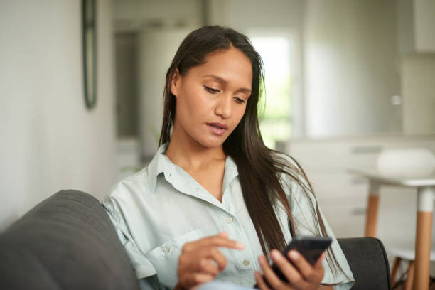 istock Tired woman using a phone inside her home looking overworked and stressed. Young indian woman browsing online and checking social media on a mobile device while sitting alone in her living room 1411569846