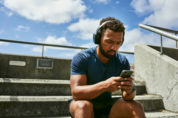 modny sportowiec ze słuchawkami słuchający muzyki na telefonie podczas relaksu na zewnątrz na stadionie. młody współczesny sportowiec robiący sobie przerwę od joggingu i cieszący się najnowszymi piosenkami za pomocą aplikacji online - spotify zdjęcia i obrazy z banku zdjęć