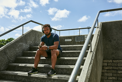Trendy runner listening to music on headphones while sitting on a staircase outside. Portrait of a young athlete taking a break from his fitness exercise to listen to songs online for motivation