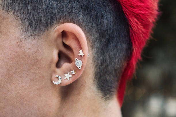 nahaufnahme des ohrs eines weiblichen punk mit silbernen piercings und einer leuchtend rot gefärbten irokesen persönlichen frisur - pierced punk goth teenager stock-fotos und bilder