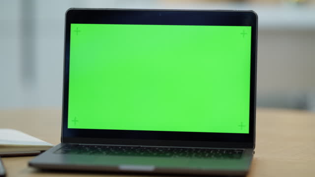 Laptop computer with mock up green screen display on table in remote home office space. Blank computer screen on wood desk, with copyspace. Closeup of bright green background display for website