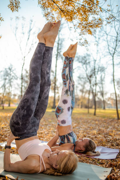 Two women doing yoga in the park in shoulder stand Candle pose Two middle aged women doing yoga on sunny autumn day outdoors, shoulder stand with legs straight up - Sarvangasana Viparita Karani, stress relief and relaxation technique shoulder stand stock pictures, royalty-free photos & images