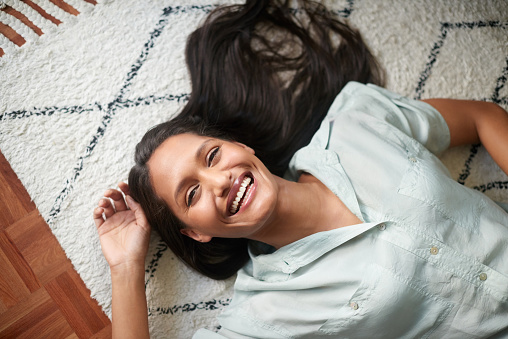 Portrait of a beautiful woman smiling and laughing with joy while lying on the floor at home from above. Cheerful, happy and carefree female enjoying a relaxing and comfortable day alone at home
