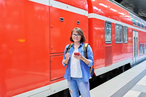 Woman passenger with smartphone waiting on railway platform inside station, smiling female with backpack looking at camera. Rail transport, passenger transportation, journey tourism travel trip people