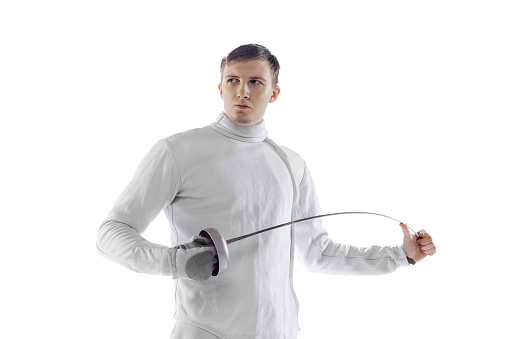 Portrait of professional male fencer in fencing costume and mask holding smallsword isolated on white background. Athlete practicing in motion, action. Copyspace for ad.