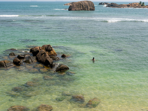Galle, Sri Lanka - March 12, 2022: Man swims in the clear water of the Indian Ocean near large stones.