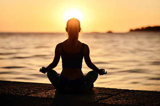 Rear view of a female athlete doing Yoga breathing exercises in Lotus position on a pier at sunset. Copy space.