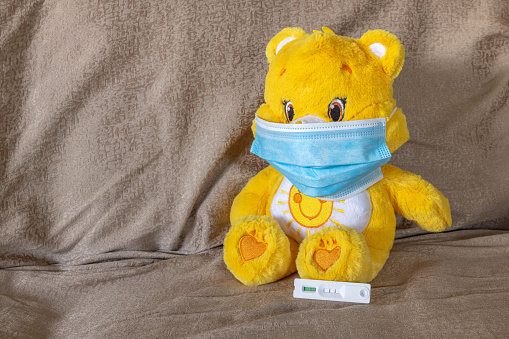 Close-up view of a yellow and white coloured teddy bear laying  on a sofa using a blue face mask during the Covid-19 pandemia with a positive rapid antigen test cassette at its feet.