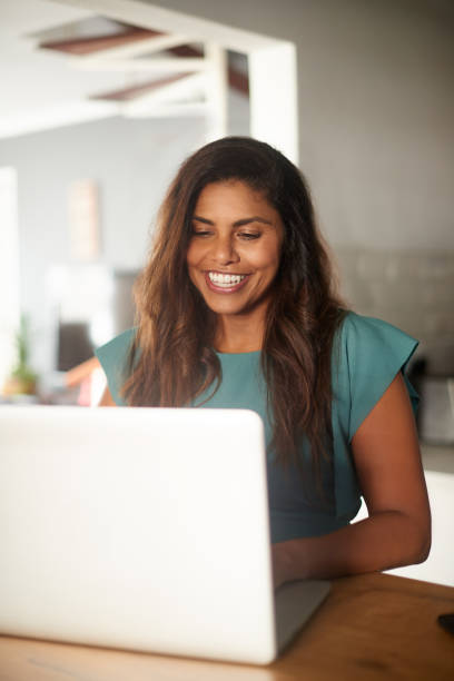 istock Happy woman using a laptop indoors. Young student doing an online assignment or studying for a virtual test inside a modern room. Cheerful entrepreneur working on a new marketing campaign from home 1411600703