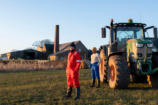 A full-length wide angle view of two proud male farmers stood with their hands in their pockets as they stand on their farm in front of a large tractor in Northumberland in thr North East of England.