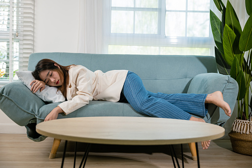 Tired woman sleeping closed eyes on sofa in living room at home after overworked working, Asian female resting falling asleep lying on couch, Full body