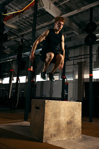 Portrait of sportive, muscular red-haired young man jumping on box, doing exercises isolated over gym background. Concept of sport, health, action, nutrition, youth. Copy space for ad