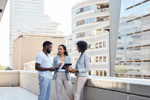 Business people talking while using a digital tablet on the balcony of an office in the city outdoors. Diverse young colleagues collaborating to discuss project plans and ideas while browsing online