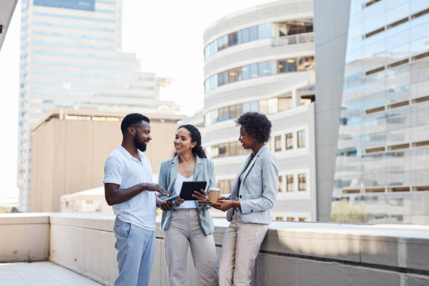 A group of professionals talking and sharing ideas for their project while standing on the office terrace with digital tablets in their hands. Adults enjoy some break time from work. stock photo