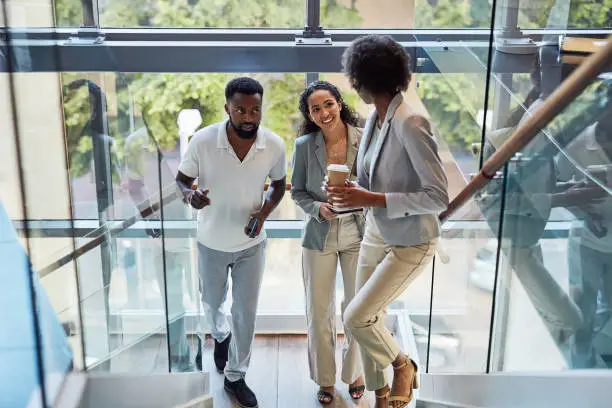 Photo of Business people walking up stairs and talking in a modern workplace. Group of young colleagues chatting on the steps of a stylish office. Coworkers on a coffee break having a discussion on staircase