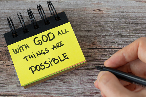 With God all things are possible, handwritten text on a yellow note with a hand on a wooden table