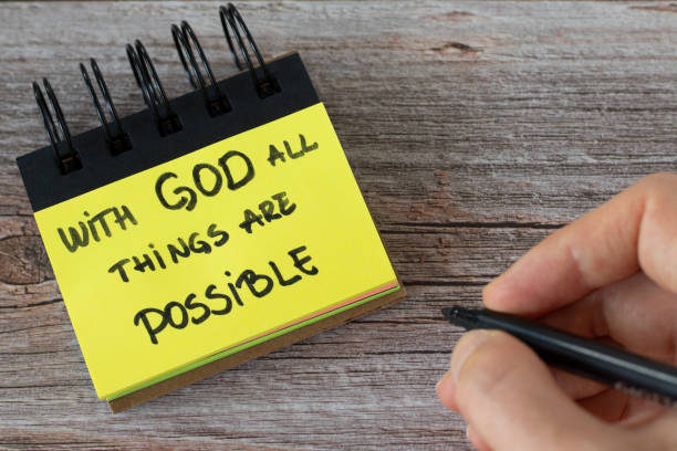 With God all things are possible, handwritten text on a yellow note with a hand on a wooden table stock photo