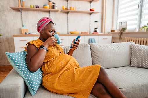 Young black beautiful pregnant woman sitting on sofa, using smart phone and drinking coffee at home.
