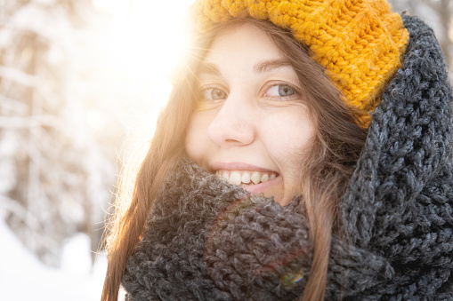 portrait of a smiling young caucasian woman in a bright woolen hat and scarf, against the backdrop of a snowy forest