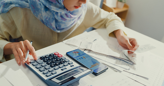 Close up Asian muslim woman with hijab using phone calculator focus on utility bills calculate check credit card receipt monthly expense bill in living room at house. Home financial lifestyle concept.