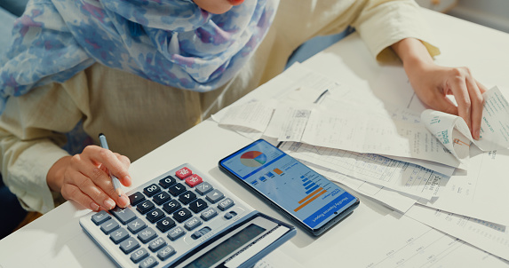 Close up Asian muslim woman with hijab using phone calculator focus on utility bills calculate check credit card receipt monthly expense bill in living room at house. Home financial lifestyle concept.