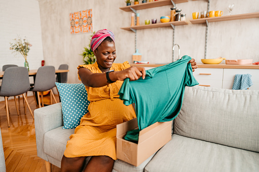 Satisfied Buyer. Portrait of pregnant black woman receiving a package, unpacking cardboard box. She is in the living room at her apartment. Happy with online purchase.