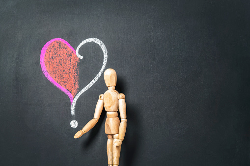 A wooden man stands next to a heart and a question mark drawn on a chalk board.