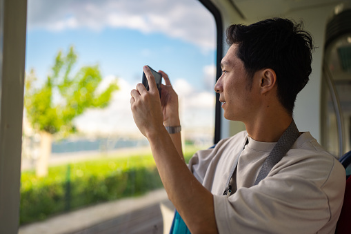 A young male tourist is riding a train and taking photos of the view from the window during his travel.