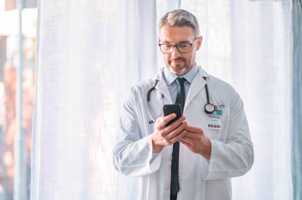 médico varón maduro usando un teléfono en una habitación de hospital moderna. un médico senior que brinda asesoramiento médico en línea sobre la aplicación de chat de comunicación confidencial. trabajador sanitario que utiliza la aplicación de te - scientist lab coat doctor male fotografías e imágenes de stock