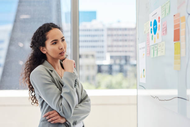istock Young business woman thinking of ideas while brainstorming and planning a project with notes on a wall in an office. Dedicated entrepreneur analyzing data and stats to develop a solution and strategy 1411625644