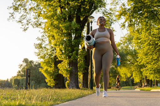 Young African American woman walking with exercise mat and bottle of water through the park in a summer day
