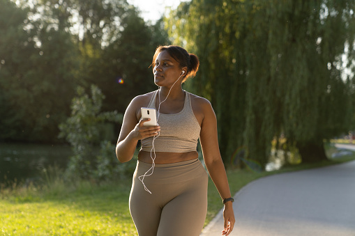 Plus sized African American woman using mobile phone and earphones during the walking at the park in a summer day